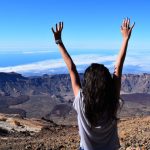 A woman celebrates atop Mount Teide with arms raised, overlooking the vast landscape and sea.
